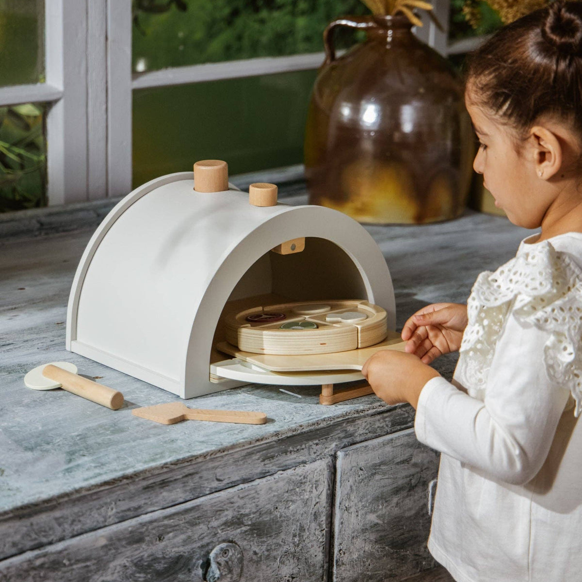 Child playing with a small white pizza oven on a wooden surface outdoors.